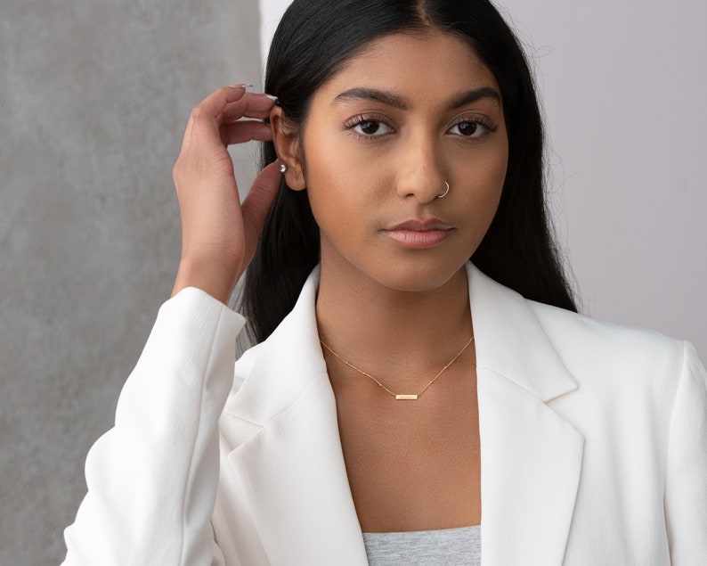 May include: A woman with long black hair wearing a white blazer and a gold necklace with a bar pendant. She is wearing a nose ring.