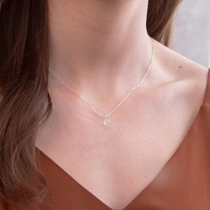 May include: A silver chain necklace with a delicate, floral pendant. The necklace is worn by a person with brown hair and a brown top.