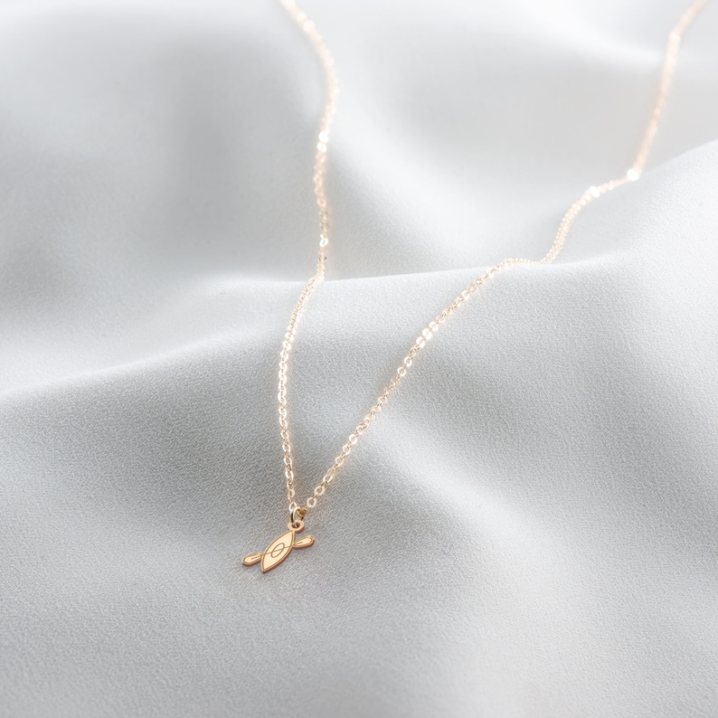 K&ouml;nnte beinhalten: Eine goldene Kettenkette mit einem kleinen goldenen Anh&auml;nger in Form eines Kanus. Die Kette liegt auf einem wei&szlig;en Stoffhintergrund.