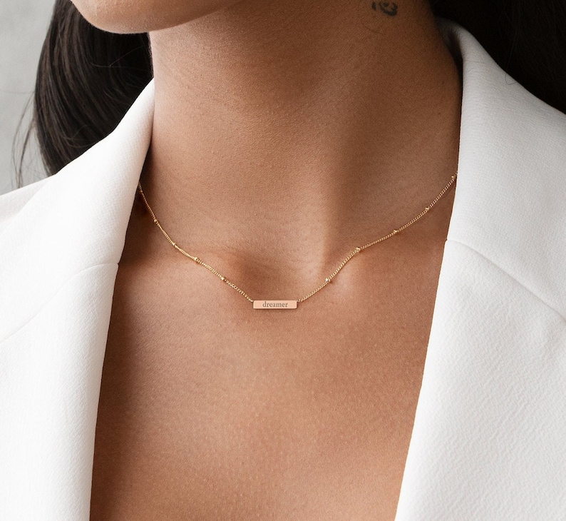 May include: A gold necklace with a delicate chain and a small bar pendant that says "dreamer".