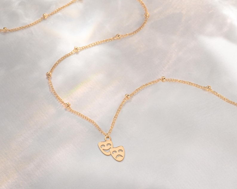 May include: A gold chain necklace with a pendant featuring two face masks, one smiling and one frowning. The chain has small gold beads.