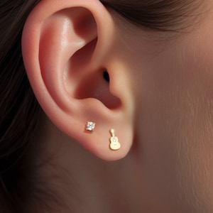 以下が含まれることがあります： 人の耳のクローズアップで、2つのイヤリングをしています。左のイヤリングは小さな透明な宝石です。右のイヤリングは金色のギターです。
