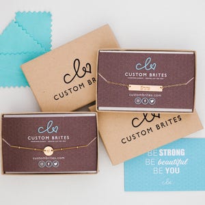 May include: Two brown boxes with the logo "clo CUSTOM BRITES" and the website address "custombrites.com". The boxes contain delicate gold necklaces with a small circle pendant and a bar pendant. A blue card with the text "BE STRONG BE beautiful BE YOU clo" is in the foreground.