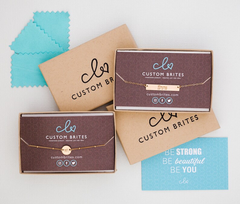 May include: Two brown boxes with the logo "clo CUSTOM BRITES" and the website address "custombrites.com". The boxes contain delicate gold necklaces with a small circle pendant and a bar pendant. A blue card with the text "BE STRONG BE beautiful BE YOU clo" is in the foreground.