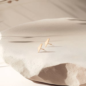 May include: A pair of gold-toned earrings shaped like the outline of a country. The earrings are on a white textured surface.