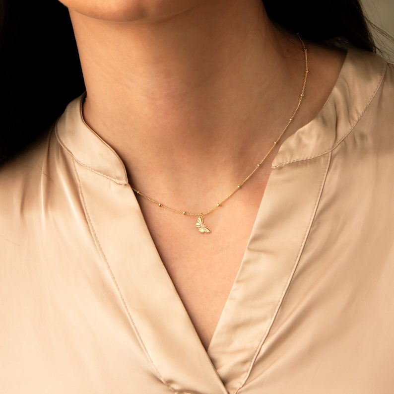 Pode incluir: Um colar de corrente de ouro com um delicado pingente de borboleta. O colar &eacute; feito de pequenas contas redondas e &eacute; usado por uma pessoa com uma camisa marrom claro.