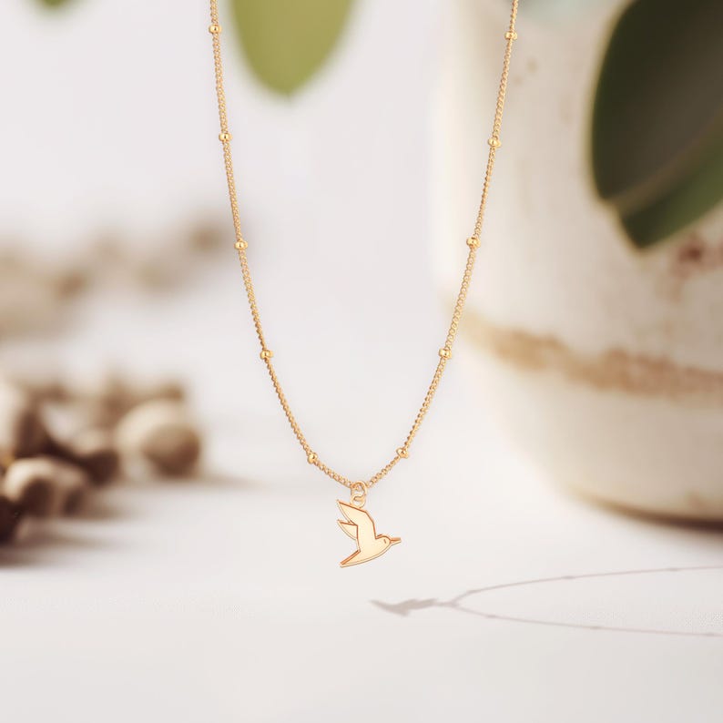 May include: A delicate gold-colored necklace featuring a small bird pendant. The chain is adorned with tiny gold beads. The bird pendant is a simple silhouette of a flying bird. The necklace is displayed against a soft, neutral background.
