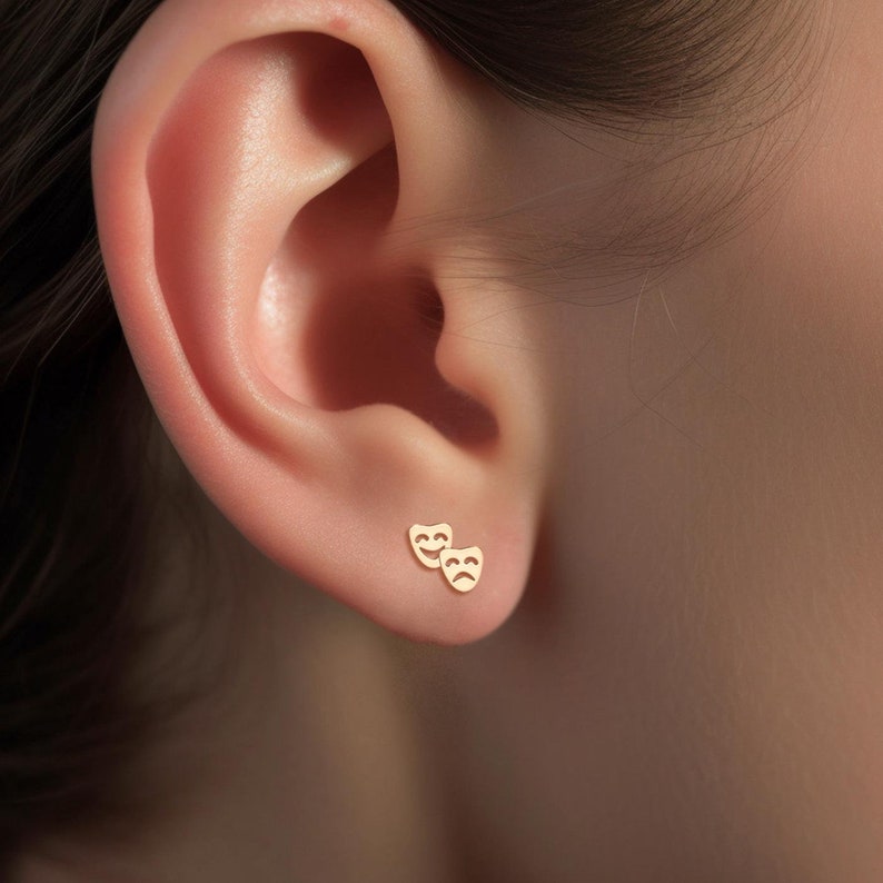 May include: A pair of gold-toned earrings featuring a smiling and a frowning face, resembling theater masks.