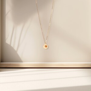 May include: A gold chain necklace with a small, round pendant featuring a sun design. The pendant is hanging against a light beige background.