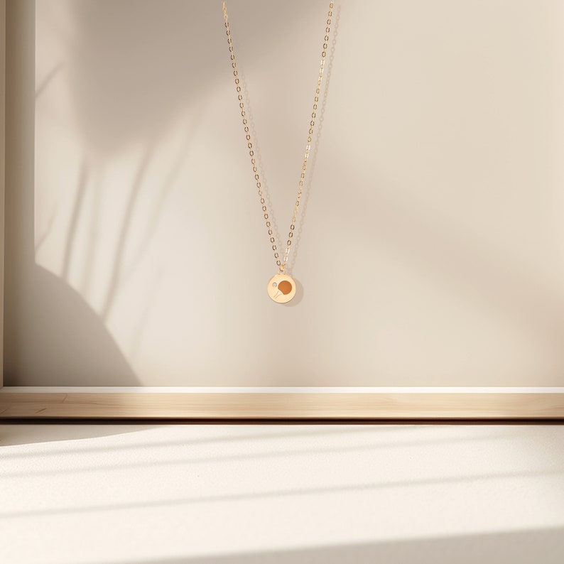 May include: A gold chain necklace with a small, round pendant featuring a sun design. The pendant is hanging against a light beige background.