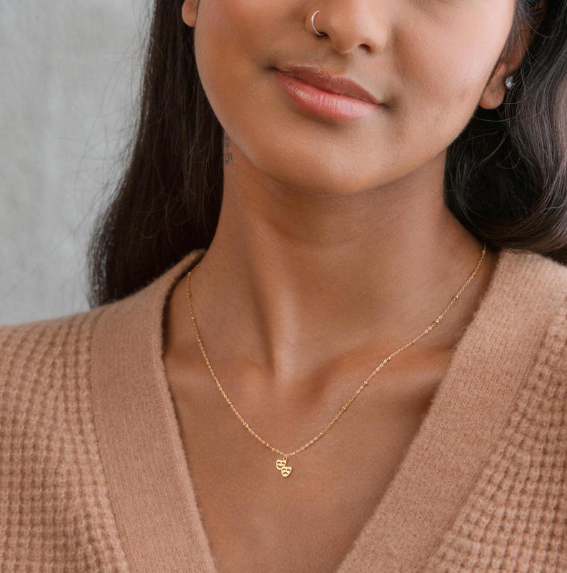 May include: A gold chain necklace with a small double heart pendant. The necklace is worn by a person with brown skin.