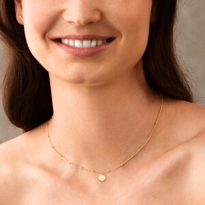 May include: A delicate gold necklace with a small, round pendant. The necklace features a fine chain with evenly spaced small beads. An arrow-shaped earring is visible in the ear. The jewelry is worn on a person.