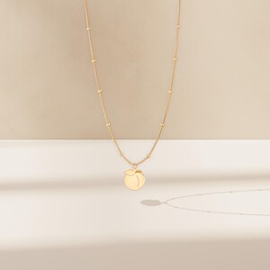 May include: A gold-coloured necklace with a peach pendant. The necklace chain features small, evenly spaced beads. The peach pendant is a simple, stylised design with a smooth surface, casting a shadow on the white surface.
