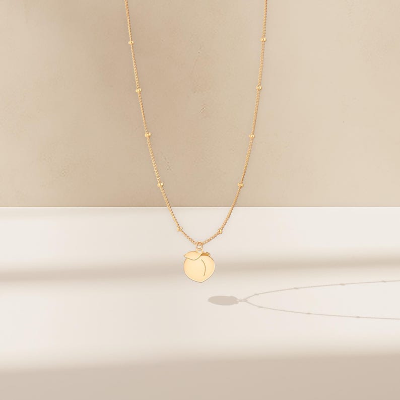May include: A gold-coloured necklace with a peach pendant. The necklace chain features small, evenly spaced beads. The peach pendant is a simple, stylised design with a smooth surface, casting a shadow on the white surface.