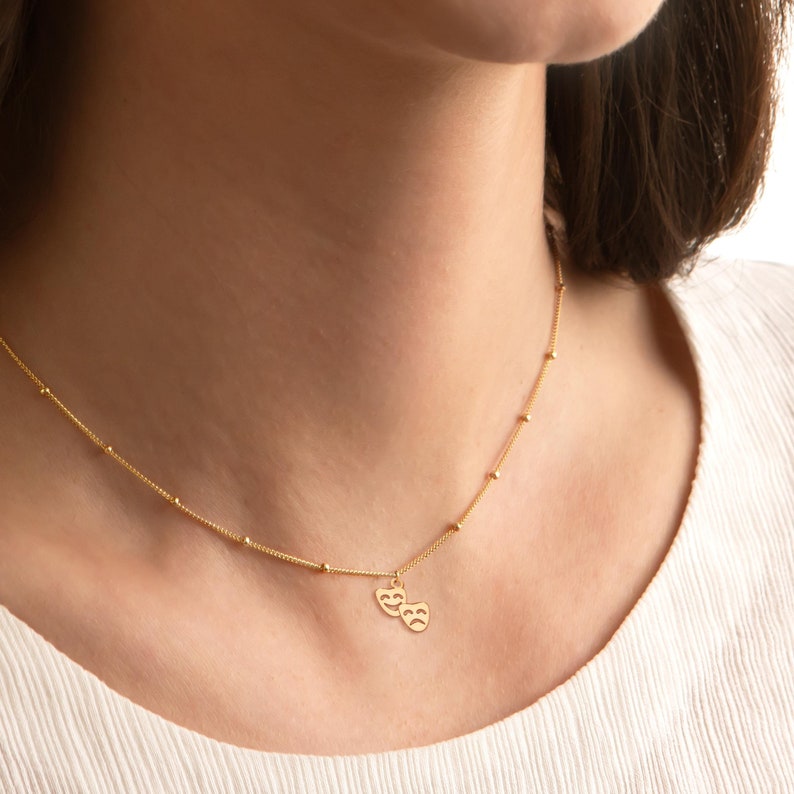 May include: A gold necklace with a delicate chain and a small pendant featuring two theater masks. The masks are facing each other, one with a smiling face and the other with a frowning face.