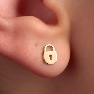 May include: A small, polished gold-tone stud earring shaped like a classic padlock. The earring features a smooth, rounded rectangular body with a curved shackle at the top and a central keyhole cutout detail, designed to sit flush against the earlobe.