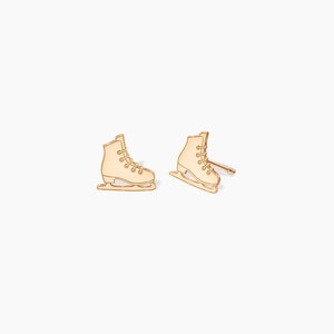 May include: A pair of gold-coloured ice skate stud earrings. The earrings are shaped like ice skates, with detailed laces and blades. The earrings are set against a white background.