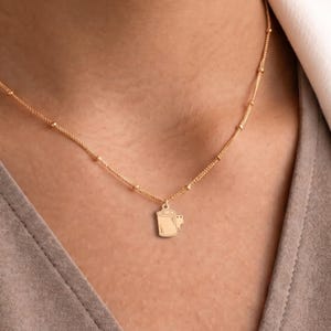 May include: A gold-toned necklace featuring a delicate chain with small evenly spaced beads and a central pendant shaped like a 35mm film canister. The pendant has a smooth surface with subtle engraved lines representing the film spool and tab.