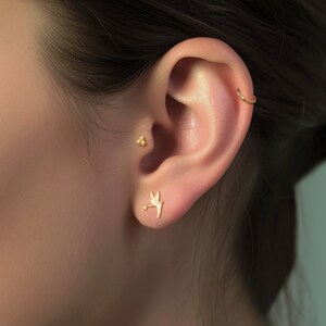 May include: A person's ear with three gold earrings. The top earring is a small cluster of three gold balls. The middle earring is a small gold fairy with wings and a wand. The bottom earring is a gold hoop.