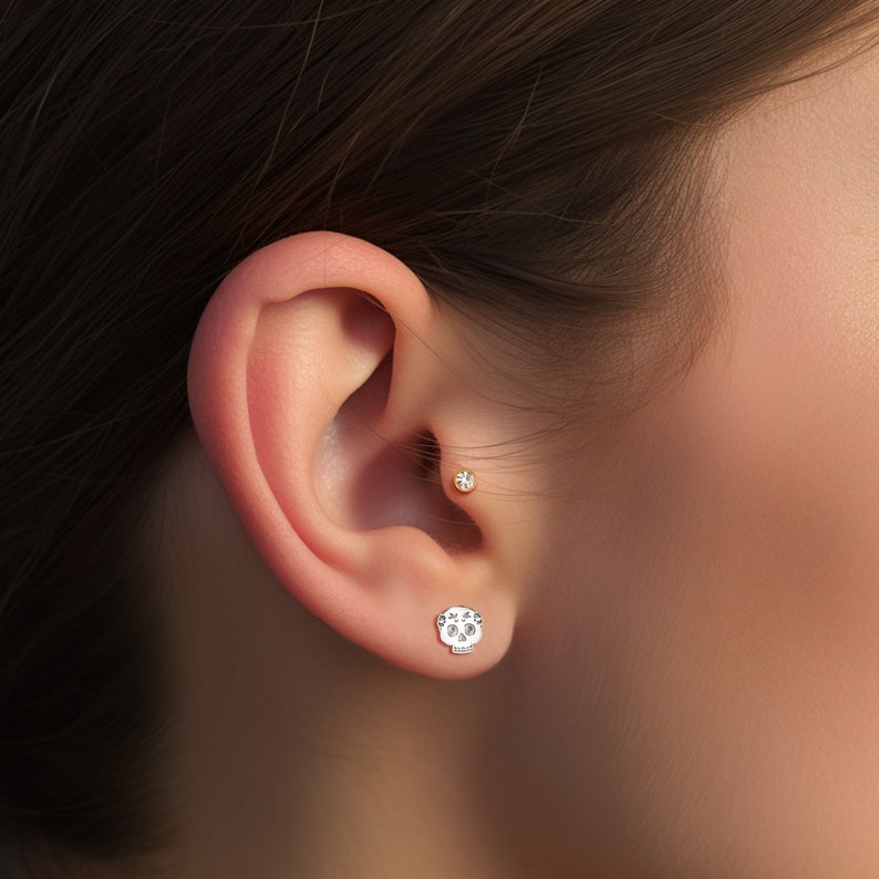 May include: A close-up of a person's ear with two earrings. The left earring is a small, round, clear gemstone. The right earring is a silver skull with small, clear gemstones for eyes.