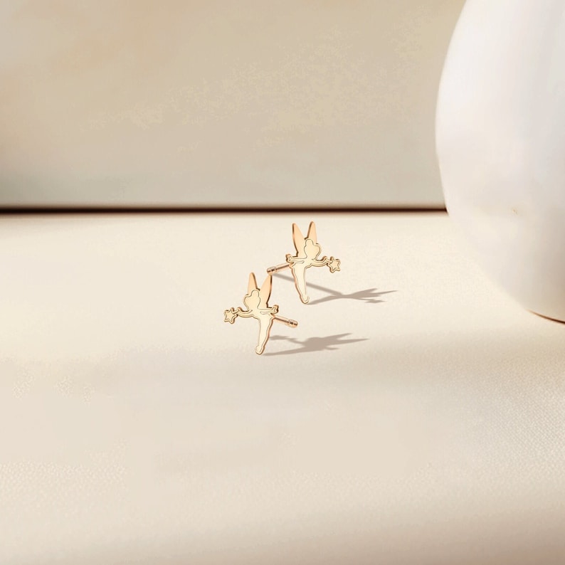 May include: A pair of gold-toned fairy stud earrings. Each earring features a delicate silhouette of a fairy with wings outstretched, holding a wand.