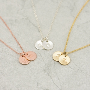 May include: Three delicate necklaces with two small round pendants each. The necklaces are in gold, silver, and rose gold. The pendants are engraved with letters and a peace sign.