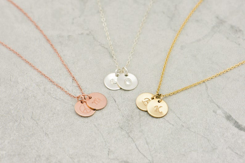 May include: Three delicate necklaces with two small round pendants each. The necklaces are in gold, silver, and rose gold. The pendants are engraved with letters and a peace sign.