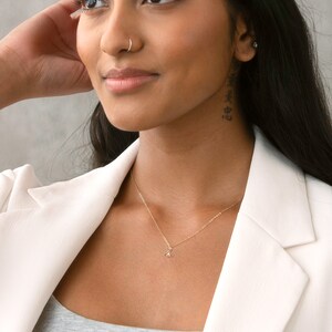 Pu&ograve; includere: Una donna indossa un blazer bianco con una collana a catena dorata con un piccolo ciondolo. Ha un piercing al naso e un tatuaggio sul collo.
