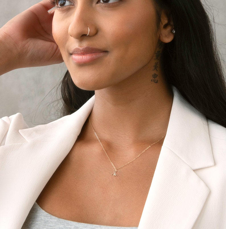 Pu&ograve; includere: Una donna indossa un blazer bianco con una collana a catena dorata con un piccolo ciondolo. Ha un piercing al naso e un tatuaggio sul collo.