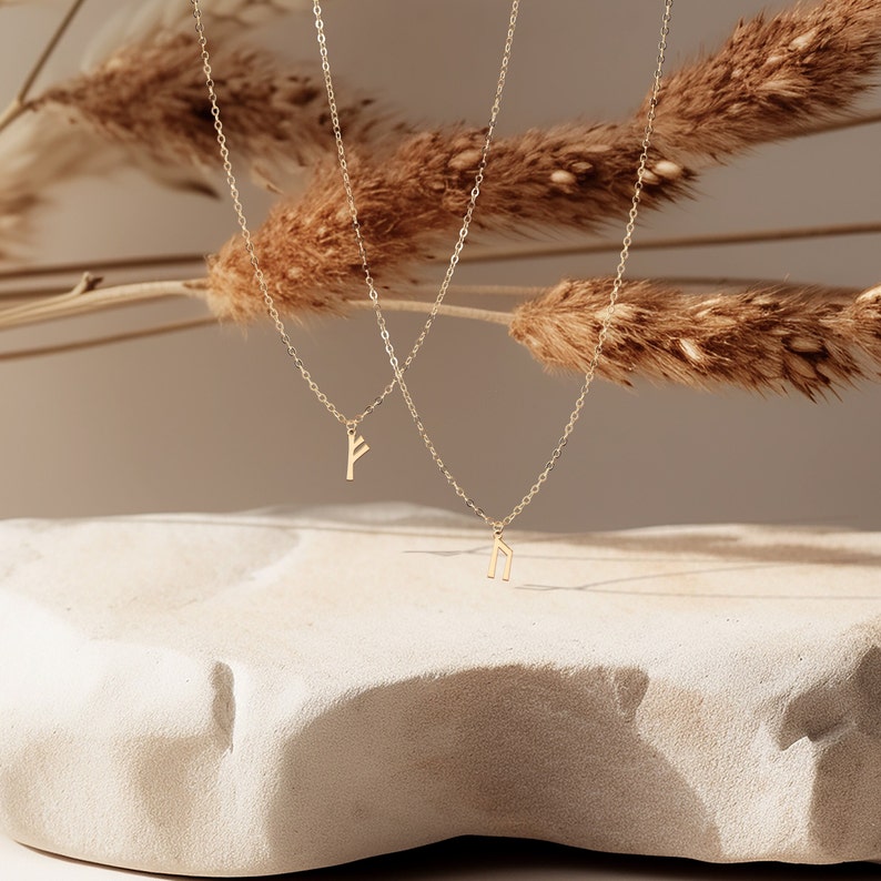 K&ouml;nnte beinhalten: Zwei goldene Halsketten mit zarten Ketten und kleinen Anh&auml;nger-Anh&auml;ngern. Die Anh&auml;nger sind in Form der Runen Algiz und Ingwaz, die Symbole aus dem Elder Futhark sind.