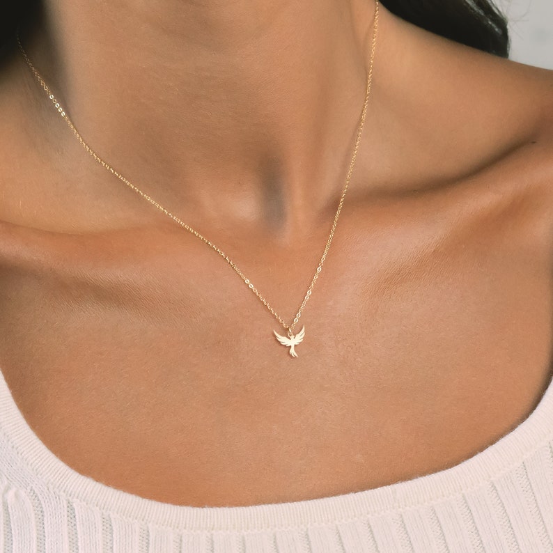 May include: A gold chain necklace with a delicate bird pendant. The bird is facing forward with its wings spread out.