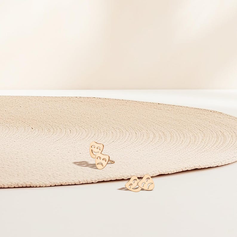 May include: A pair of gold-toned earrings featuring a smiling and a frowning face, resembling theatrical masks. The earrings are on a light beige surface.