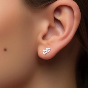 May include: A pair of silver earrings with a simple design of two theater masks, one smiling and one frowning.