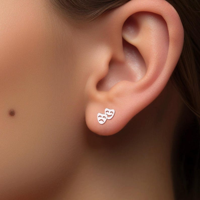 May include: A pair of silver earrings with a simple design of two theater masks, one smiling and one frowning.