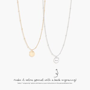 May include: Two necklaces with fine chains and small bead details. One is gold with a peach pendant, the other is silver with a round pendant engraved with the name "Oliver". The image includes text that reads "make it extra special with a back engraving!"