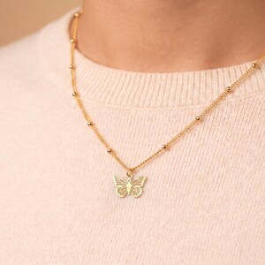 May include: A gold-toned necklace featuring a delicate chain with evenly spaced small round beads and a central butterfly pendant. The butterfly charm has a cut-out, filigree-style wing design, creating a light and airy appearance on the metallic surface.