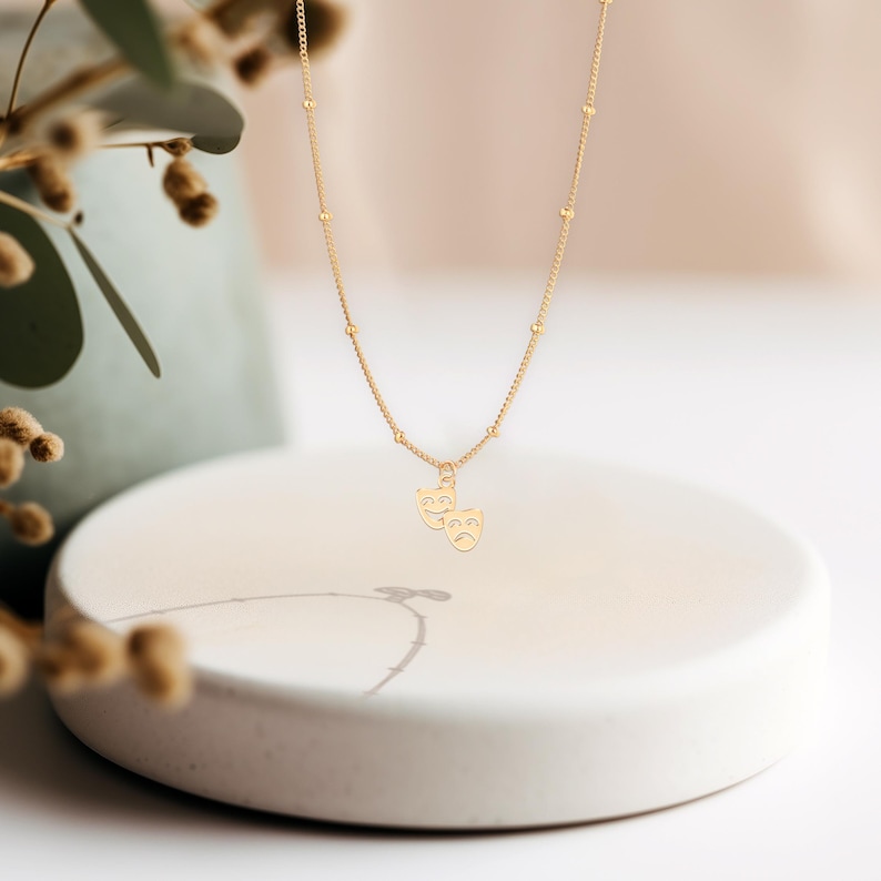 May include: A gold necklace with a delicate chain and a pendant featuring two theater masks, one smiling and one frowning.