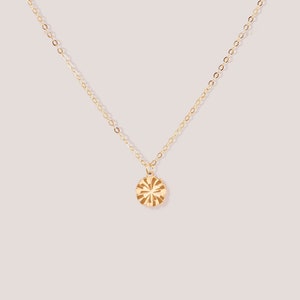 May include: A delicate gold necklace with a circular pendant. The pendant features a sunburst design with etched lines radiating from the center. The chain and pendant have a polished, shiny finish, set against a soft pink background.