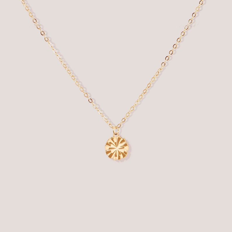 May include: A delicate gold necklace with a circular pendant. The pendant features a sunburst design with etched lines radiating from the center. The chain and pendant have a polished, shiny finish, set against a soft pink background.