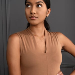 May include: A woman wearing a brown sleeveless top with a gold chain necklace and a small pendant.