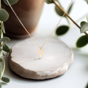 May include: A gold necklace with a delicate female symbol pendant. The necklace is displayed on a light gray stone surface.