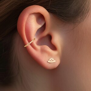 May include: An ear featuring gold jewellery. A slender ear cuff curves around the upper ear, and a small stud earring shaped like a UFO is in the lobe. The jewellery has a warm, golden colour.