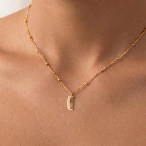 May include: A gold-toned necklace featuring a delicate chain with evenly spaced small spherical beads and a central pendant shaped like a miniature comb. The metal has a polished finish, and the design is suspended from a thin, refined link chain.