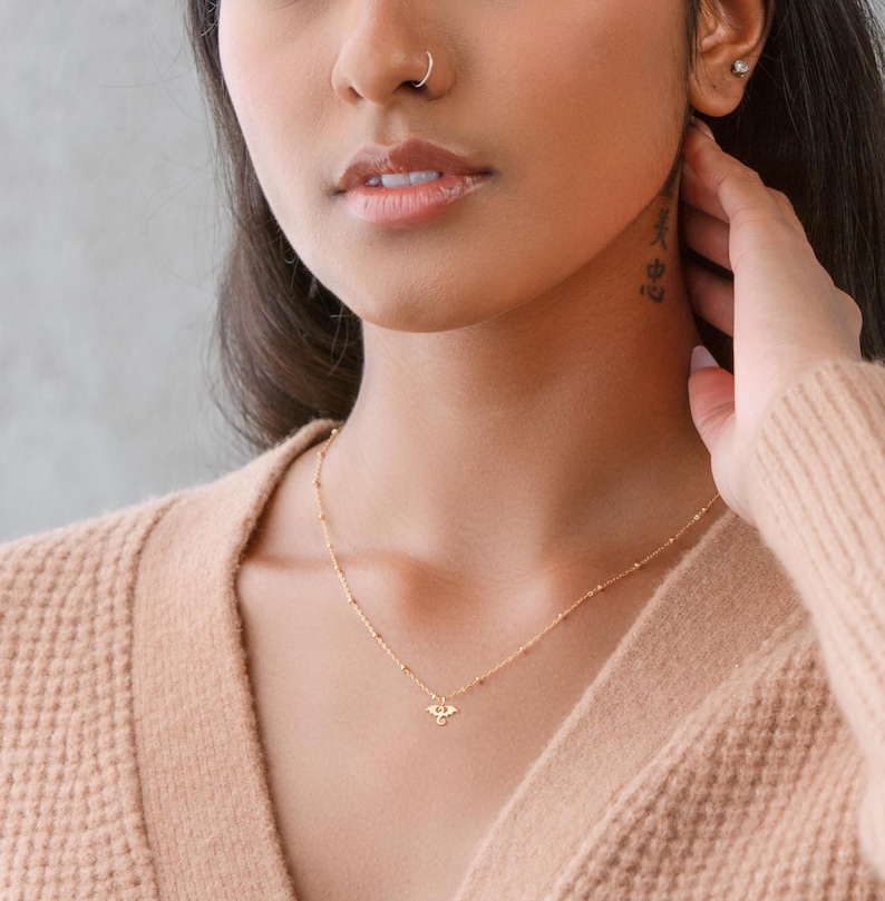 May include: A gold chain necklace with a small eye pendant. The necklace is worn by a person with a nose ring and a tattoo on their neck.