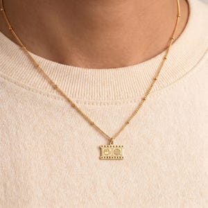 May include: A gold-toned necklace featuring a delicate chain with small evenly spaced beads and a rectangular pendant shaped like a film strip. The pendant displays two engraved icons: a vintage movie camera and a circular film reel.