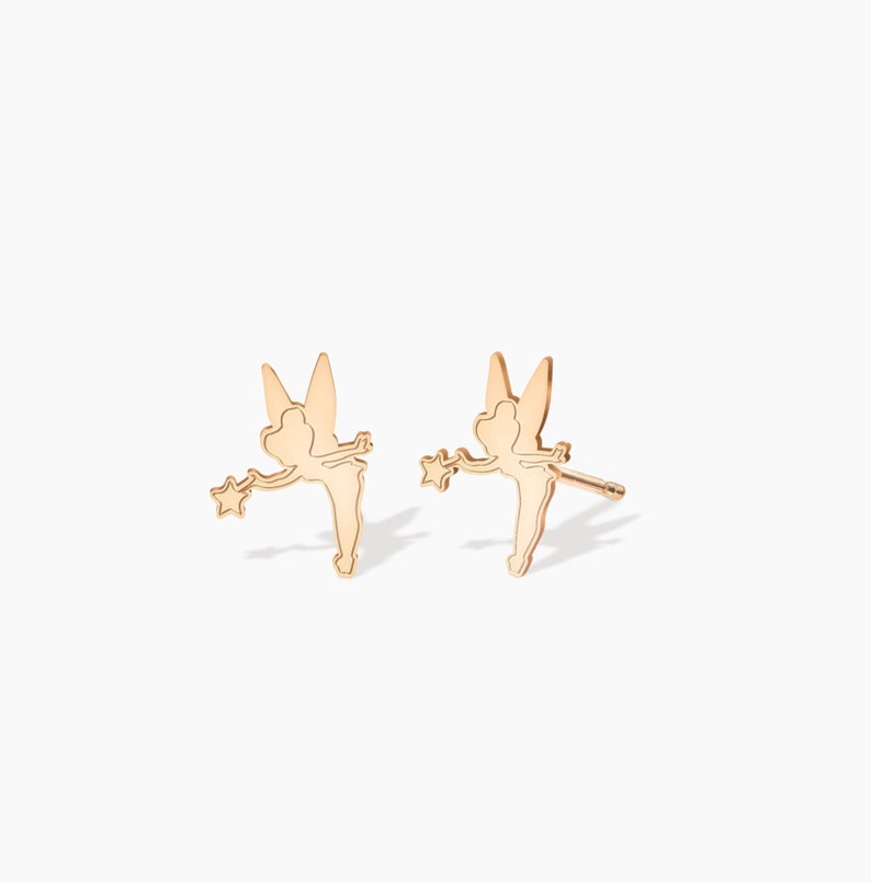 May include: A pair of gold-toned fairy stud earrings. Each earring features a delicate silhouette of a fairy with wings outstretched, holding a wand with a star at the end.