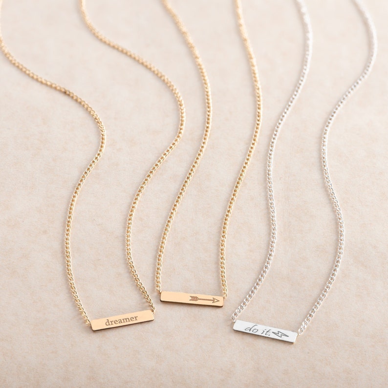 May include: Three gold and silver necklaces with delicate chains and small rectangular pendants. The pendants have the words "dreamer" and "do it." engraved on them, along with a small arrow.
