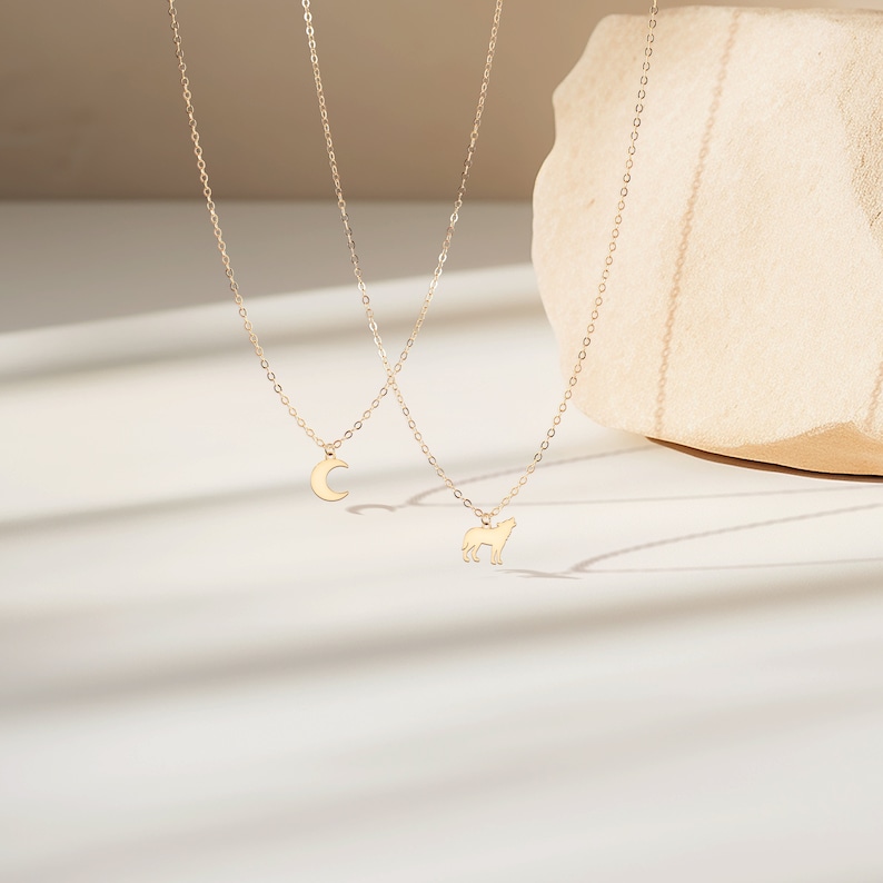 May include: Two gold necklaces with delicate chains. One necklace features a crescent moon pendant, while the other has a wolf silhouette pendant.