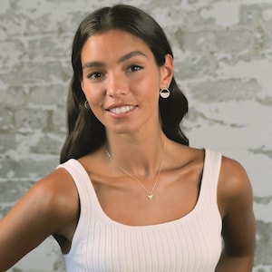 May include: A woman wearing a white tank top with a delicate gold necklace featuring a small bird pendant. She is wearing gold hoop earrings with a textured design.