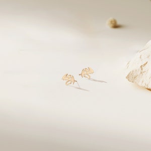 May include: A pair of gold dragon stud earrings. The earrings are small and delicate, and the dragons are facing forward with their wings spread out.